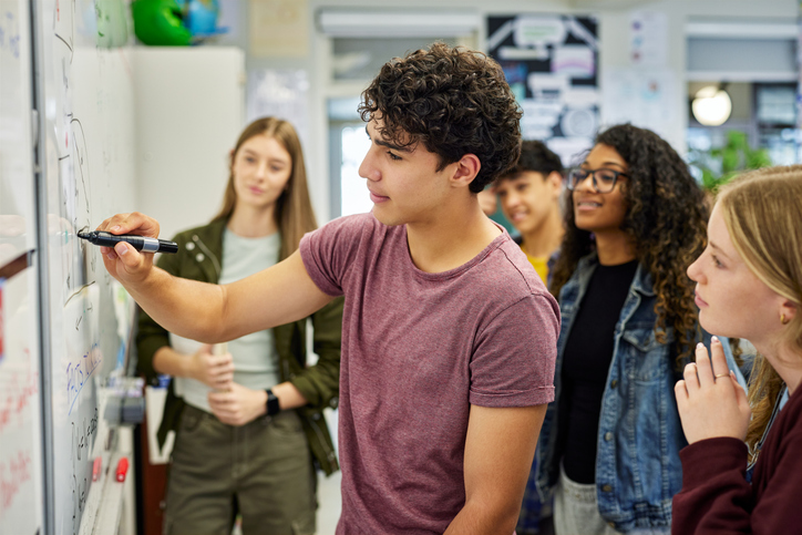 Un élève de lycée explique un concept à ses camarades en écrivant au tableau. Des adolescents concentrés présentent un exposé de groupe tout en dessinant au tableau, sous le regard attentif de leurs camarades. Un jeune garçon aide ses camarades à comprendre la solution d'un problème de géométrie ou d'arithmétique en classe.