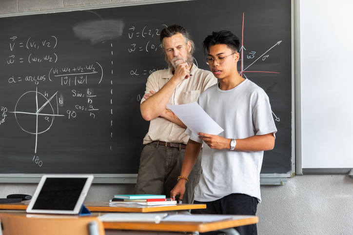 Un lycéen fait un exposé en classe tandis qu'un professeur observe ses notes.
