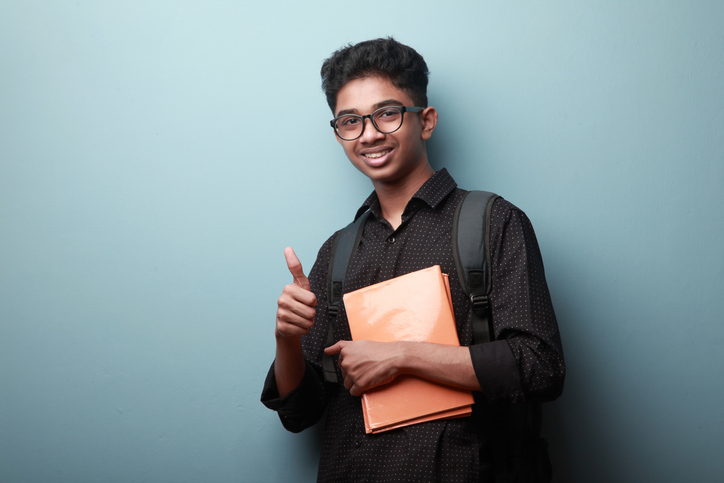 Lycéen souriant d'origine indienne tient des cahiers et fait un signe de pouce levé.