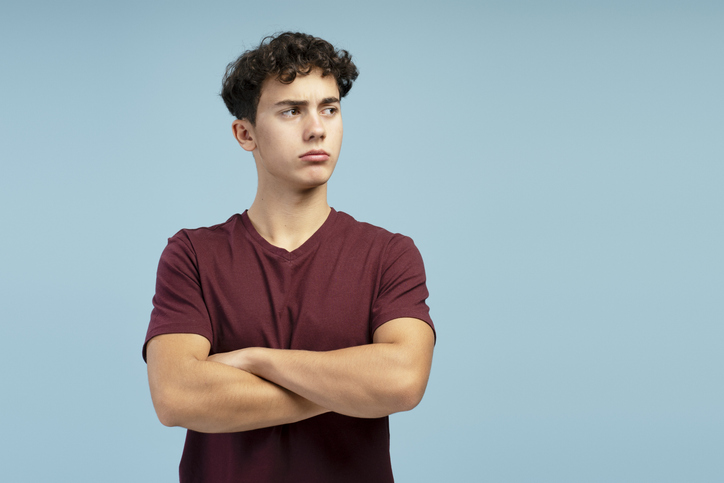 Adolescent sérieux et triste, les bras croisés, détournant le regard vers un espace libre, debout sur fond bleu.