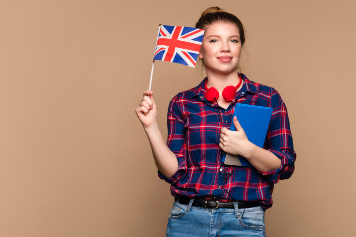 Une étudiante tient un petit drapeau britannique et un cahier dans un studio sur fond beige. Cours particulier d'anglais. Apprentissage de l'anglais britannique.