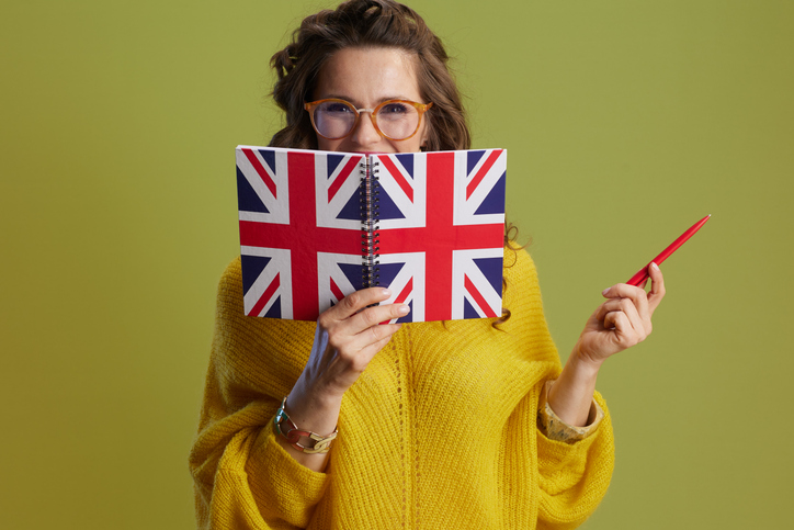 Une professeur d'anglais souriante, portant un pull jaune et des lunettes, pose sur fond vert avec un carnet aux couleurs du drapeau britannique.