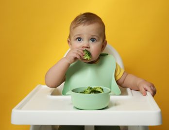 Adorable petit bébé portant un bavoir mange des brocolis sur fond jaune.