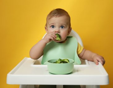 Adorable petit bébé portant un bavoir mange des brocolis sur fond jaune.