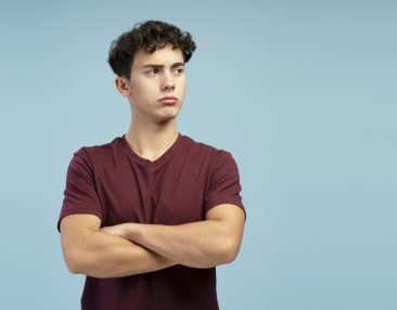 Adolescent sérieux et triste, les bras croisés, détournant le regard vers un espace libre, debout sur fond bleu.