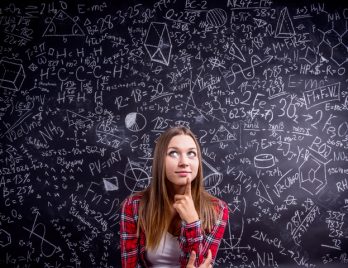 Etudiante de première au lycée à Thionville en chemise à carreaux rouges se tient devant un grand tableau noir couvert de symboles et de formules mathématiques d'équations du second degré.