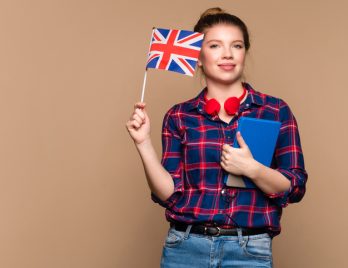 Une étudiante tient un petit drapeau britannique et un cahier dans un studio sur fond beige. Cours particulier d'anglais. Apprentissage de l'anglais britannique.
