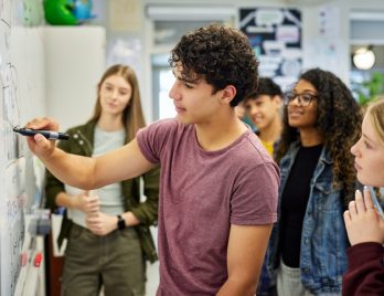 Un élève de lycée explique un concept à ses camarades en écrivant au tableau. Des adolescents concentrés présentent un exposé de groupe tout en dessinant au tableau, sous le regard attentif de leurs camarades. Un jeune garçon aide ses camarades à comprendre la solution d'un problème de géométrie ou d'arithmétique en classe.
